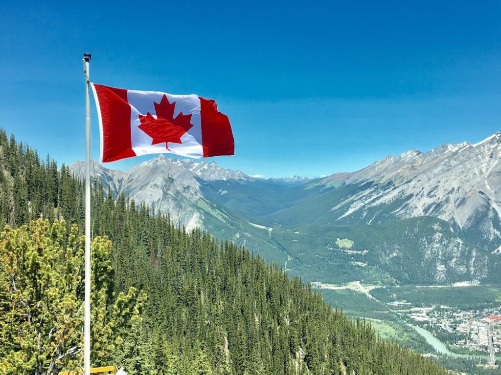 zipwp-image-756790 Canadian flag waving atop a scenic mountain view with blue skies and lush greenery.